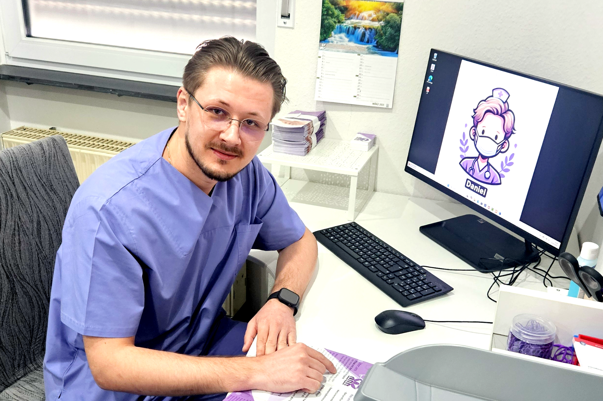 Mann in lilaner Pflegeruniform sitzt an einem Schreibtisch mit einem Computer und Grafiken.